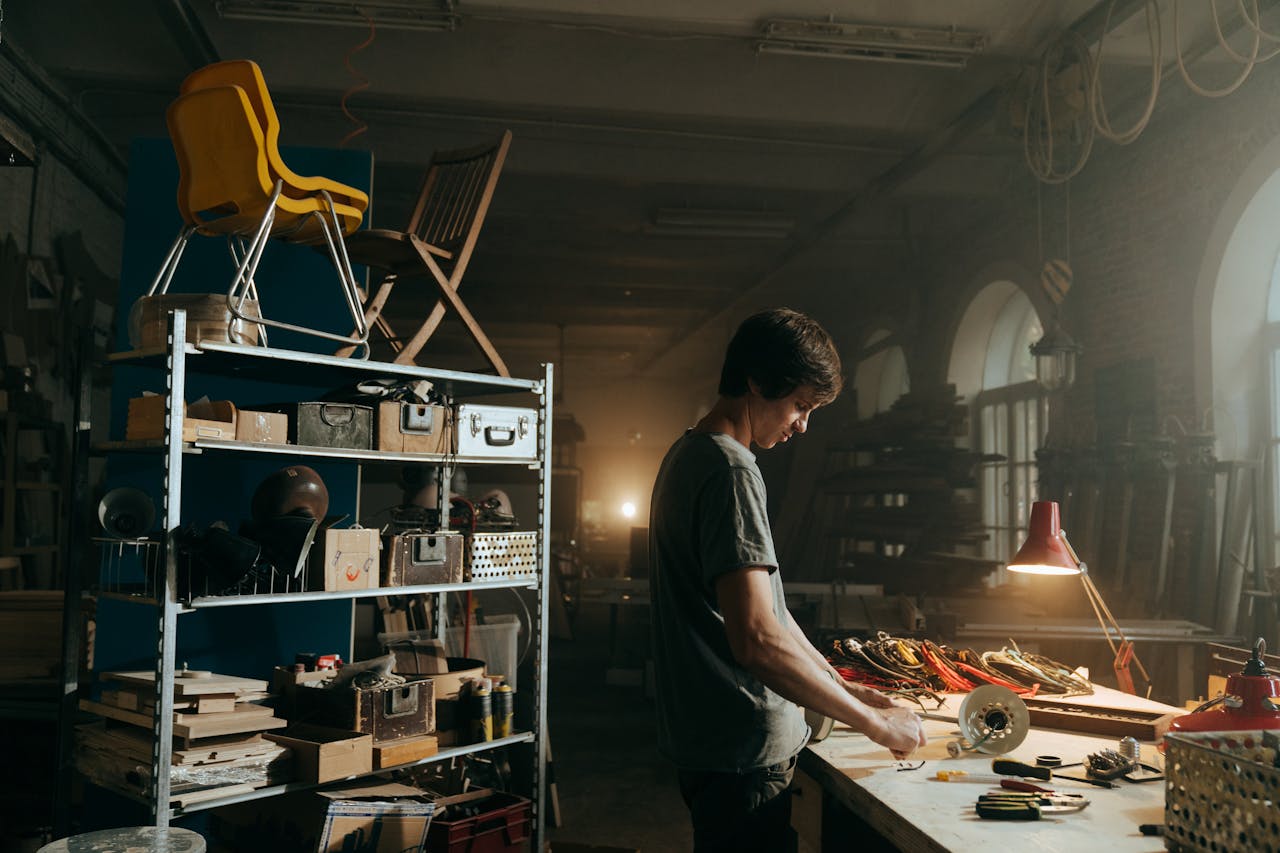 A male craftsman focuses on a project in a dimly lit vintage workshop surrounded by tools and furniture.